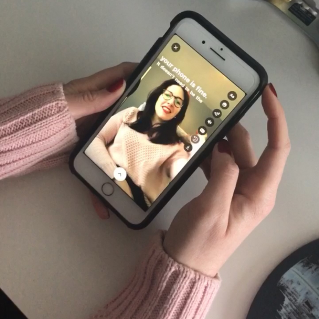 a woman's hands are holding a smartphone, the screen is unlocked and is showing an image of a woman with dark hair, glasses who is wearing red lipstick and smiling. There are closed captions on the screen