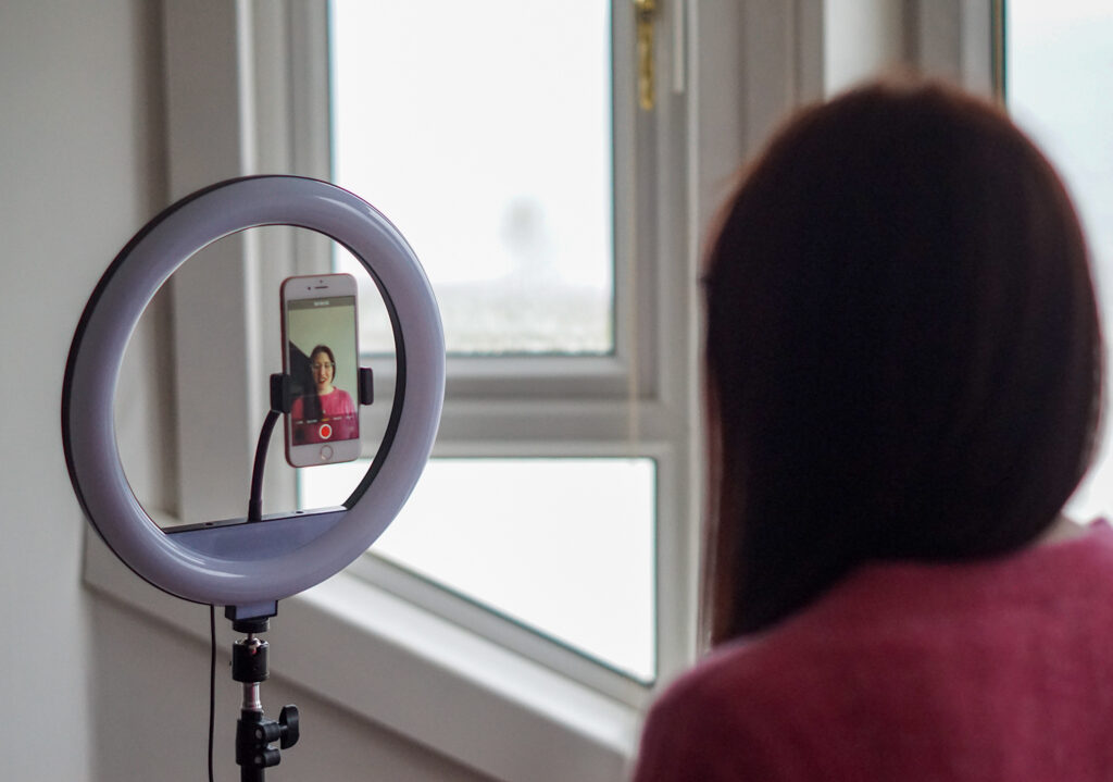 a smartphone is attached to a ring light, the screen is unlocked and is showing an image of a woman with dark hair, glasses who is wearing red lipstick and a pink jumper and smiling. The back of the woman's head is also in the shot, the photo is being taken from behind her